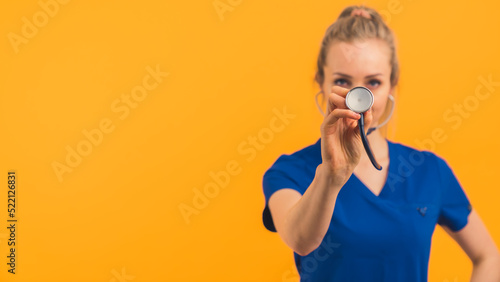 Caucasian blond female doctor in scrubs standing with hand on hip holding stethoscope up to camera. Horizontal studio shot. High quality photo