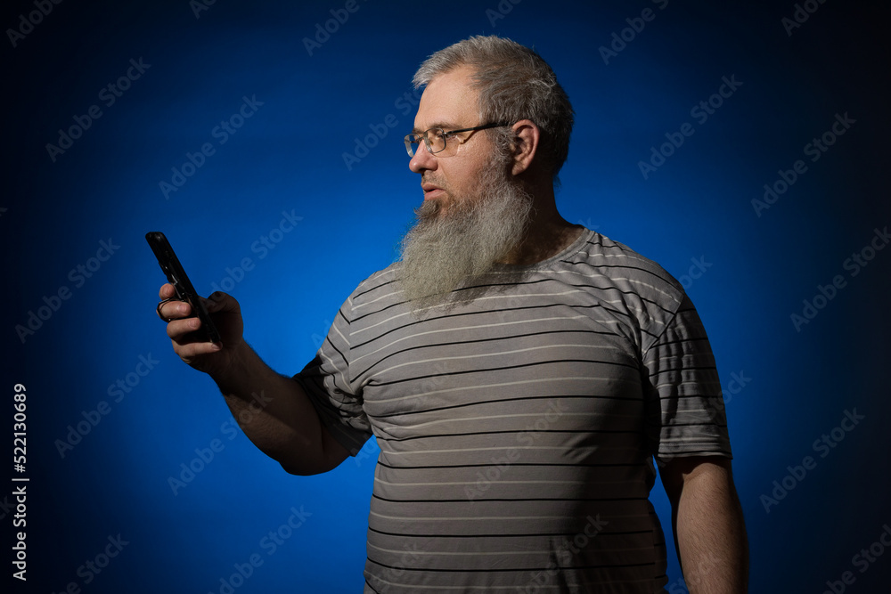 A gray-haired, bearded, disheveled, adult man in glasses and a striped shirt, with a phone in his hand. He looks at his phone and swipes. Cellular communication concept. On a blue background. A gray-haired, bearded, disheveled, adult man in glasses and a striped shirt, with a phone in his hand. He looks at his phone and swipes. Cellular communication concept. On a blue background.