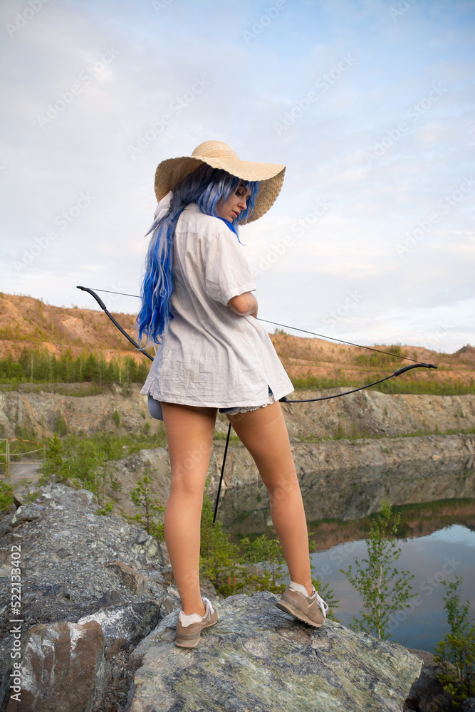 a beautiful girl in a blue wig and a straw hat with a bow and arrow in her hands.beautiful view of the lake and mountains.the concept of the game