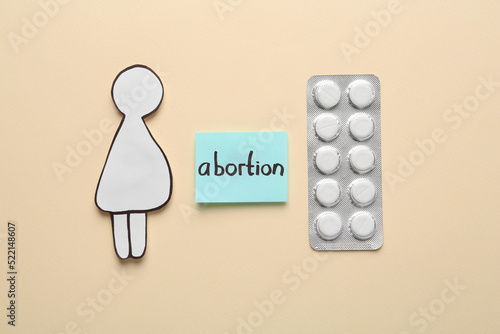 Note with word Abortion, pills and pregnant woman paper cutout on beige background, flat lay photo