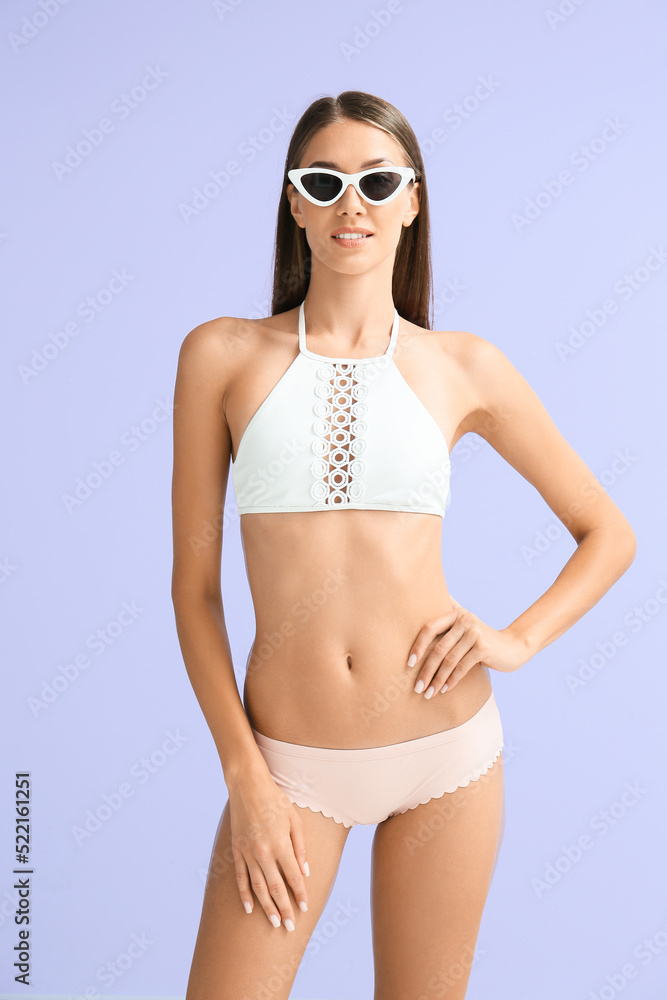 Fototapeta premium Young tanned woman in swimsuit and sunglasses on lilac background