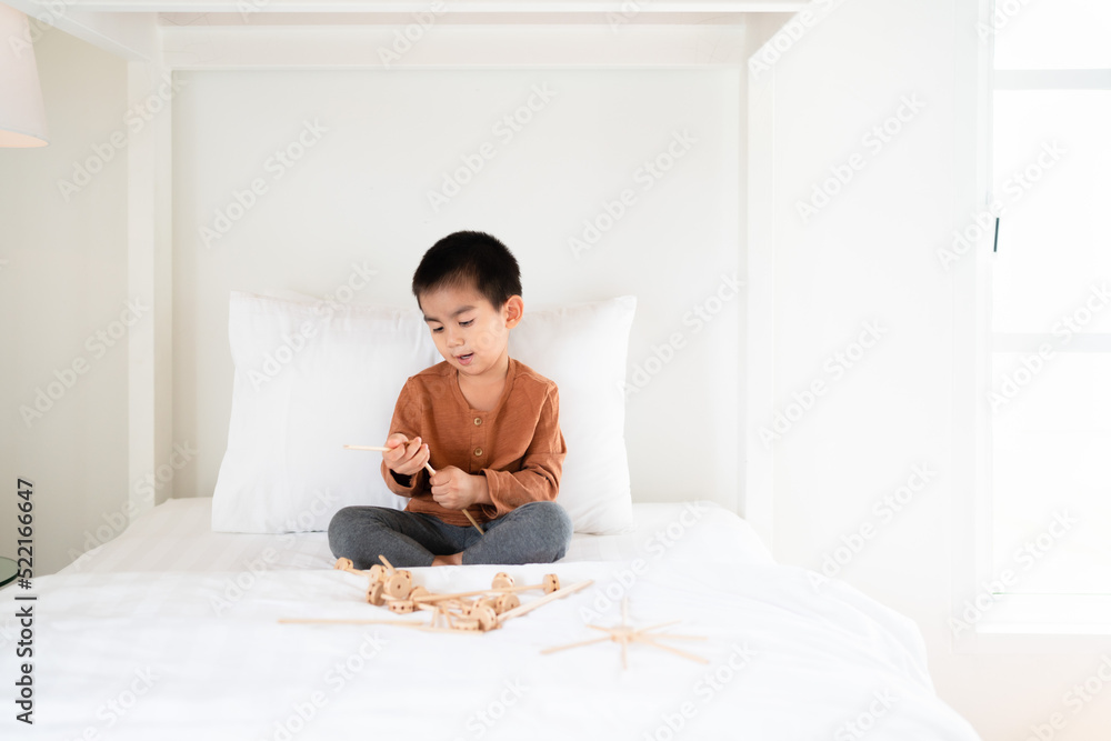 Fototapeta premium Asian little boy is playing wooden toys on the bed in the morning time, concept of learning, homeschool, education, creativity, steam, stem, robotic. Activity for kid development.