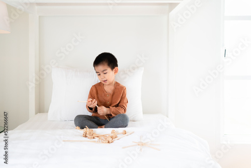 Asian little boy is playing wooden toys on the bed in the morning time, concept of learning, homeschool, education, creativity, steam, stem, robotic. Activity for kid development.