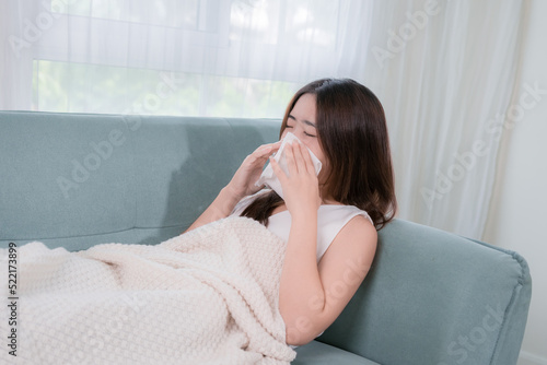 Asian woman patient with tissue her sick symptom in living room at home, Sick young female sitting under the blanket on sofa coughing or sneezing in tissue.