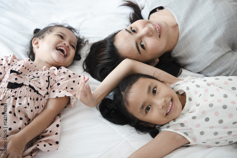 Top view of cute little girl and her beautiful sister with mother looking at camera and smiling while lying on the bed at home. Soft focus Top view of cute little girl and her beautiful sister with mother looking at camera and smiling while lying on the bed at home. Soft focus