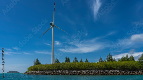 The wind generator is located on an island near the ocean. Blades on a blue sky background. Green vegetation on the ground. Seychelles