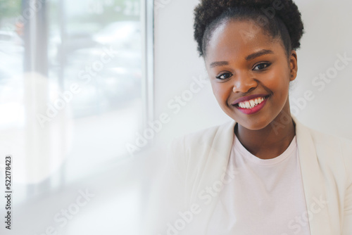 Afro woman in white jacket posing smiling at camera standing in the office. Successful and confident. Close up portrait.