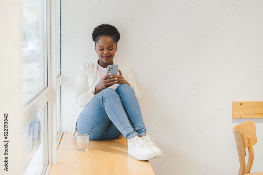 African woman sitting on the windowsill of a cafe, writing messages to her customers. Checking email. Electronic wireless device concept