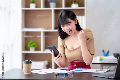 Beautiful Asian businesswoman is excited about her success, raising her hand with her mobile phone in the office.