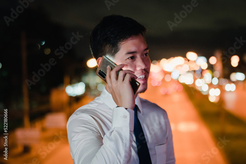 Asian businessman use mobile phone, smart phone after work walking through city street while waiting car in the city at night