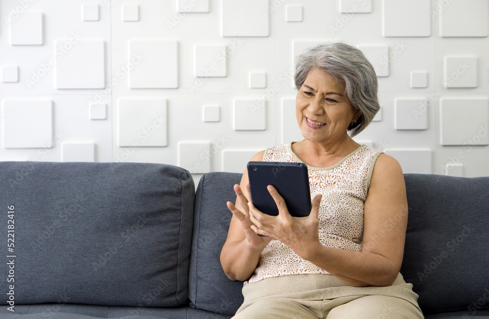 Asian gray hair old woman looking at tablet computer monitor, smiling while sit on sofa in the living room. Asian gray hair old woman looking at tablet computer monitor, smiling while sit on sofa in the living room.