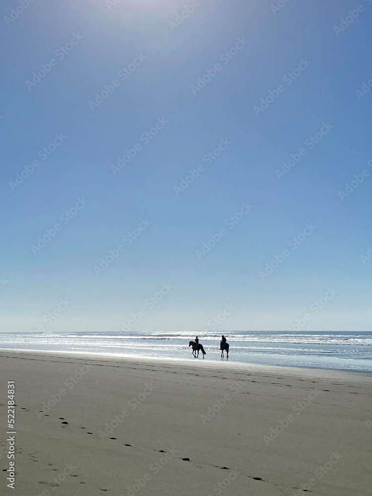 Looking into the sun at two horseback riders on a coastline beach. Blue sky in the background.