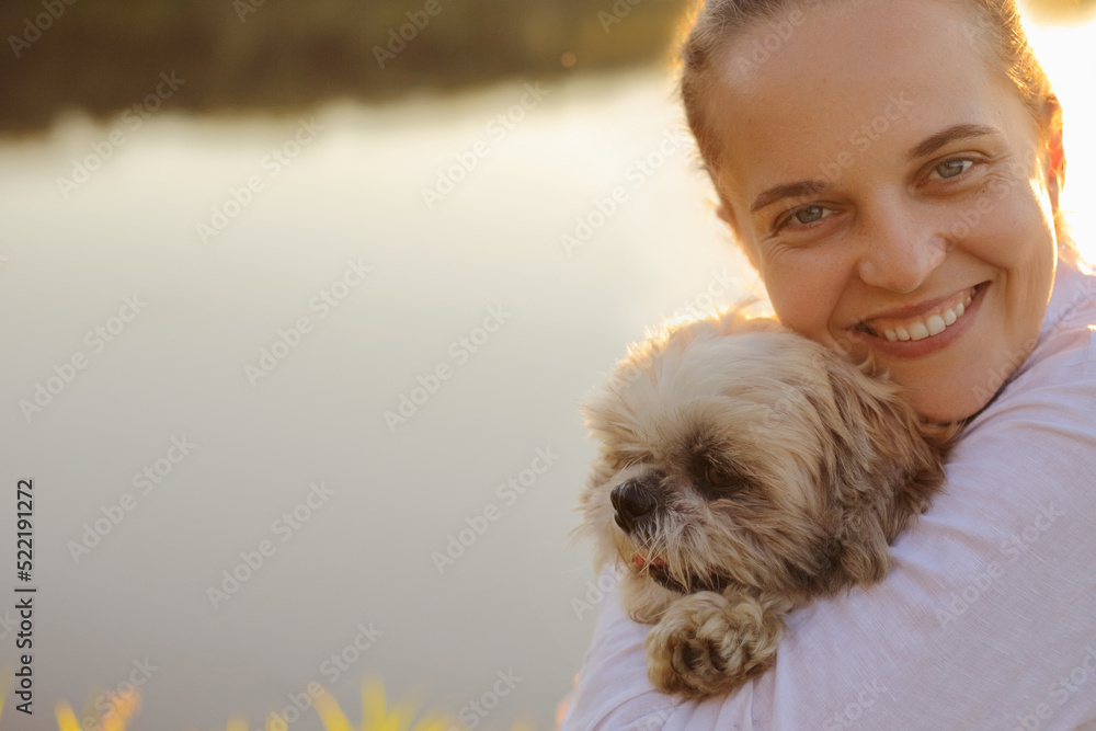 Closeup portrait of happy satisfied charming young adult woman wearing white shirt and her Pekingese dog sitting at sunset near the river, hugging her pet and looking at camera.