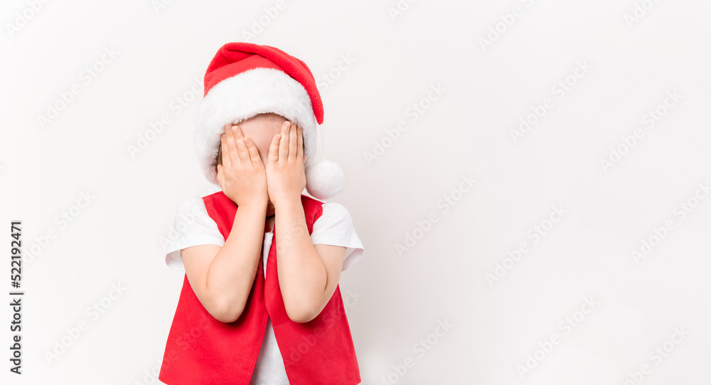 Portrait of a child covering his face with his hands in a suit and hat of Santa Claus isolated on white background. Hide and seek game. 5 year European boy. Banner. Copy space. Peek a boo. Surprise Portrait of a child covering his face with his hands in a suit and hat of Santa Claus isolated on white background. Hide and seek game. 5 year European boy. Banner. Copy space. Peek a boo. Surprise