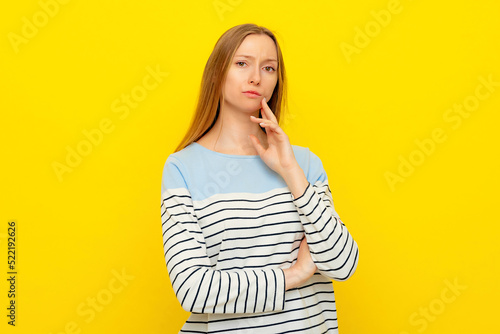 Portrait of thoughtful female, keeps finger on cheek, contemplates about future wedding with boyfriend or making important decision, dressed in casual striped sweatshirt, yellow background