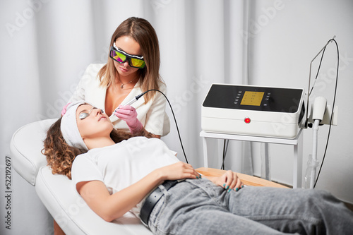 Modern treatment of telangiectasias on skin. Beautician performing local high-precision removal of dilated small vessels on woman face with innovative yellow laser. photo