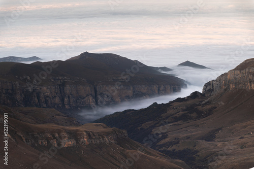 Mountains in the clouds at sunset. Aerial view of a mountainous rocky range in the fog. Beautiful landscape with high cliffs and sky. Top view from a drone of a mountain valley in low clouds