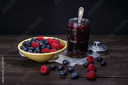 Blueberries and raspberries in a yellow bowl next to a jar of jam on a lacy linen napkin. A few berries are scattered on a wooden table