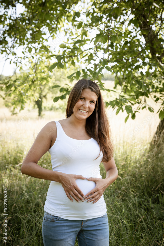 pretty attractive young pregnant woman with white top is standing in high meadow and forming a heart with her hands on her belly out in the nature pretty attractive young pregnant woman with white top is standing in high meadow and forming a heart with her hands on her belly out in the nature
