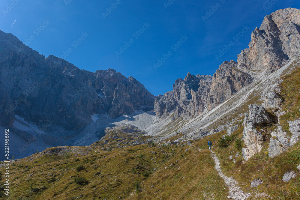 Path at the foots of rocky ridges of Croda Rossa di Sesto and Cima Undici Mountains in Comelico region with lonely mountaineer in the middle of green meadows, Dolomites, Italy