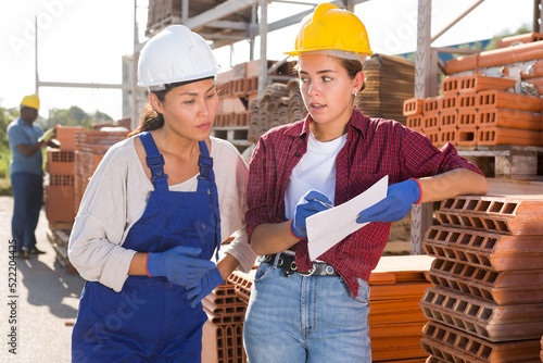 Asian and European women in hardhats discussing documentation while standing in outdoor construction material storage. African-american man standing in background.