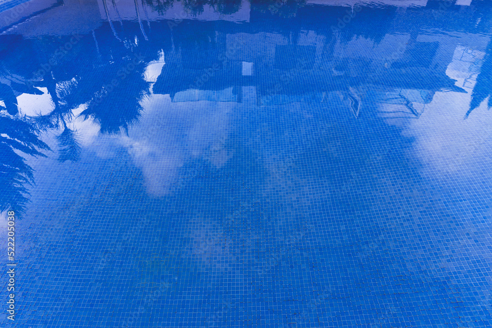 Obraz premium Swimming pool with blue water on a sunny day. Houses and palm trees are reflected in the water. The tile is visible through a transparent clean aod. July in summer. Spain.