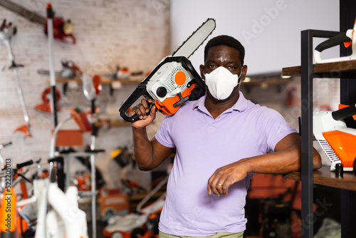 African-american man in face mask standing in salesroom of gardening tools shop with chainsaw in hand.