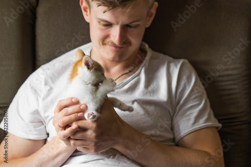 Young caucasian man in white t-shirt holding with hands a small white and red kitten at home. Man loves his pet.