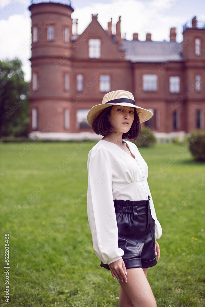 portrait of a beautiful young woman in a hat and a white blouse and shorts goes on a green meadow in summer against the background of a large castle building.
