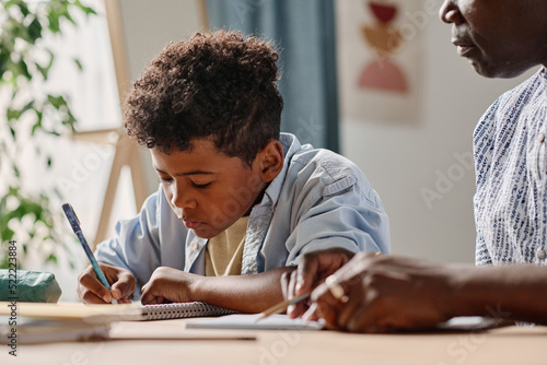 African little boy making notes in notebook while sitting at table together with teacher at home