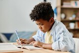 African schoolboy sitting at his desk in the room and doing homework making notes in notebook