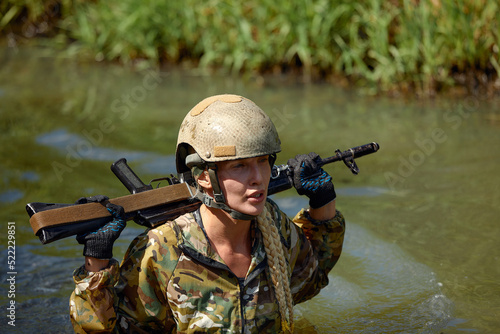 Portrait of fit military female holding weapon rifle in hands in river, posing alone. Confident strong athlete lady looking at side seriously. Attractive good-looking female in military gear