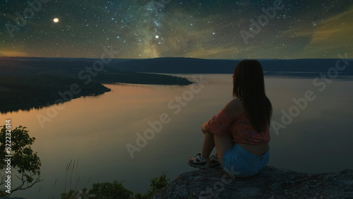 The girl sitting on the mountain top above scenic river on starry sky background