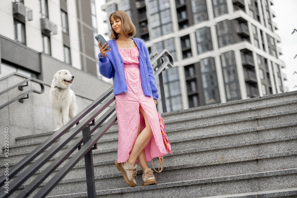 Young stylish woman dressed brightly stands with a smart phone on staircase at modern housing estate outdoors. Concept of using gadgets at everyday life Young stylish woman dressed brightly stands with a smart phone on staircase at modern housing estate outdoors. Concept of using gadgets at everyday life