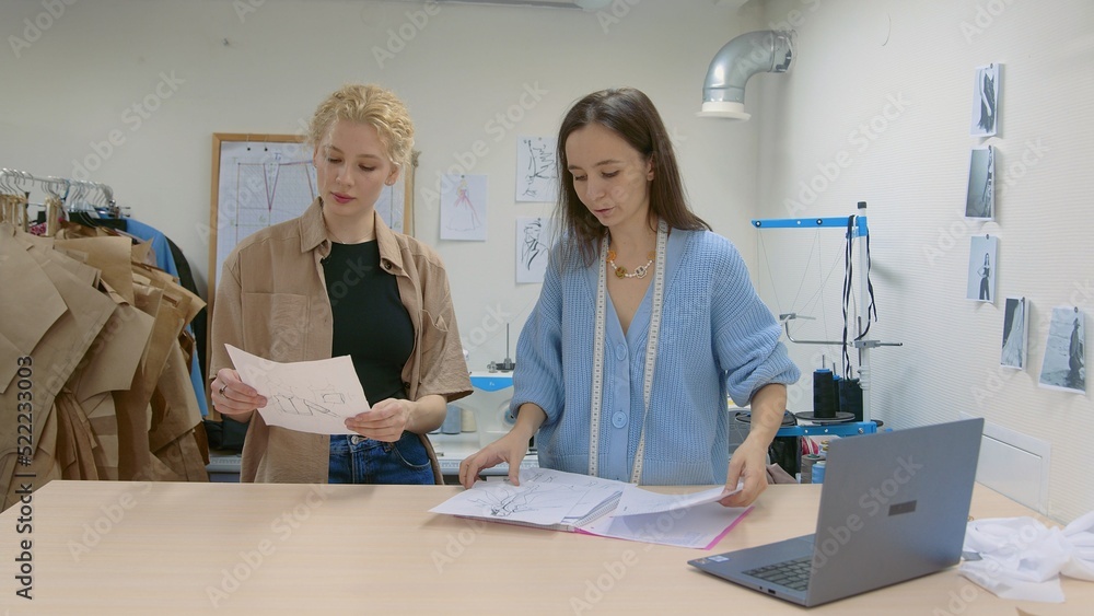 4K. A young female seamstress works with a client in a sewing workshop. They are looking at sketches of dresses 4K. A young female seamstress works with a client in a sewing workshop. They are looking at sketches of dresses