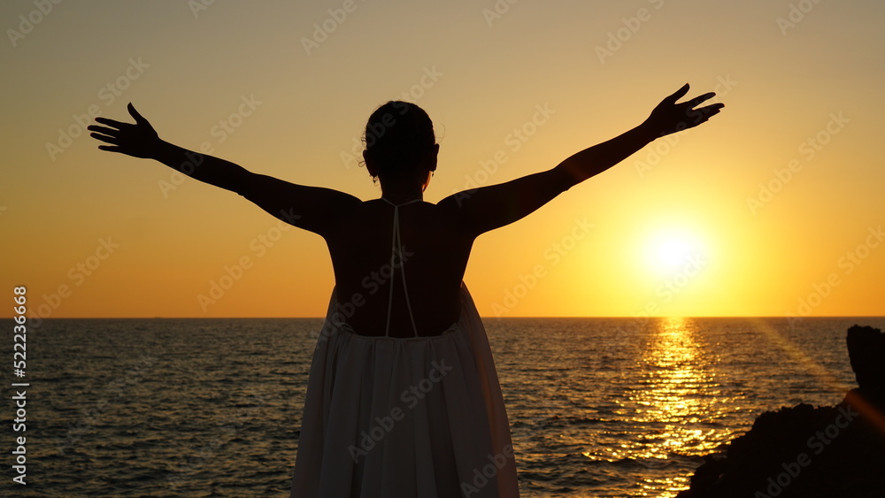 Elegant woman facing the sunset on the beach, she opens her arms holding a hat, she feels free and powerful