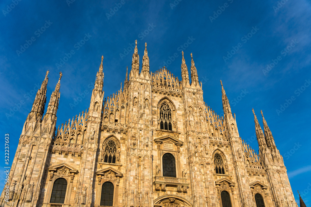 Fototapeta premium Sunset Scene of The ornate gothic facade, soaring spires and magnificent marble towers of the Duomo, Milan's monumental cathedral under big blue Lombardy skies