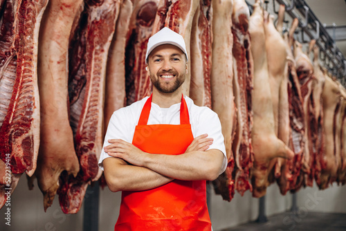 Fotografi Young butcher at the meat factory standing by the hanging meat