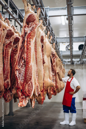 Obraz na plátně Young butcher at the meat factory standing by the hanging meat