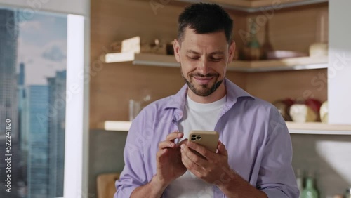 Portrait of handsome young Caucasian man with beard holding smartphone in hands in kitchen. City view from window. Communication concept. Gadget, mobile phone, technology. Indoors