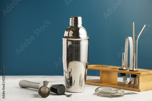 Bartender equipment - stainless steel silver colored cocktail shaker, hawthorne strainer, pourers, bar spoon, muddler , in a bamboos stand on white table on blue background photo