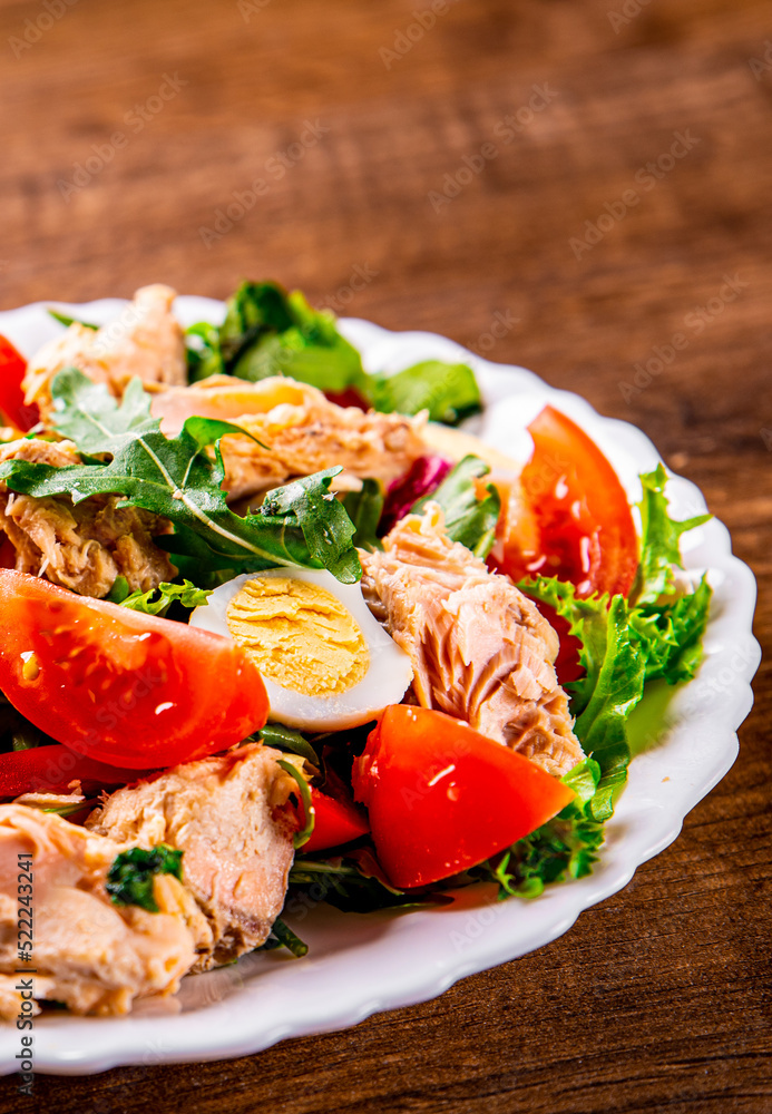 Fresh salad with fish, arugula, eggs,red pepper, lettuce, fresh sald leaves and tomato on a white plate on wooden table background - obrazy, fototapety, plakaty 