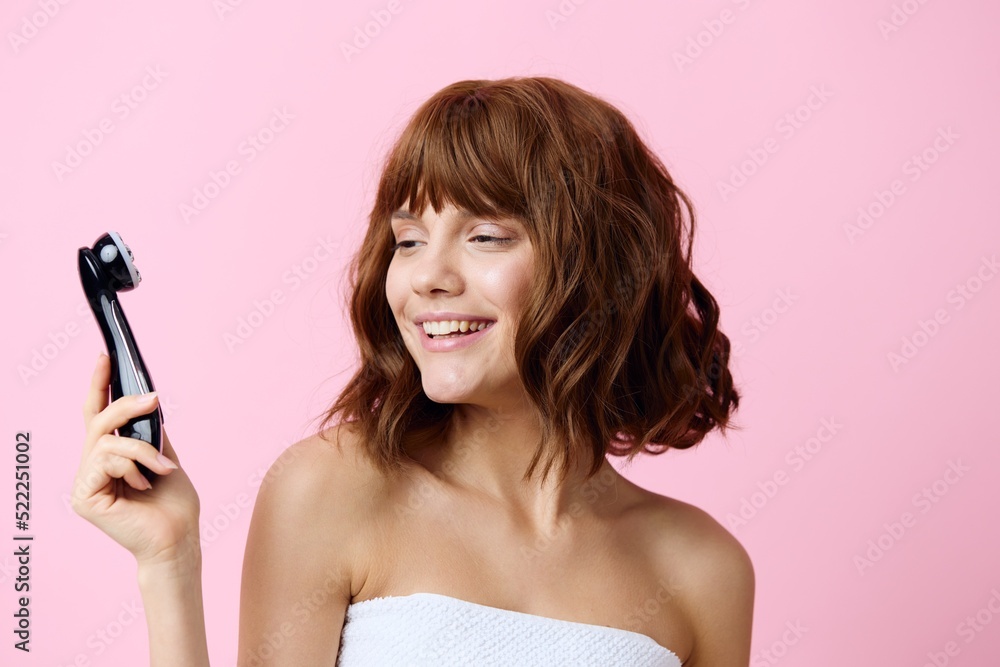 a happy, joyful, young woman stands with beautifully styled hair on a pink background and holding a black electric massager in her hand, looks at him with admiration a happy, joyful, young woman stands with beautifully styled hair on a pink background and holding a black electric massager in her hand, looks at him with admiration