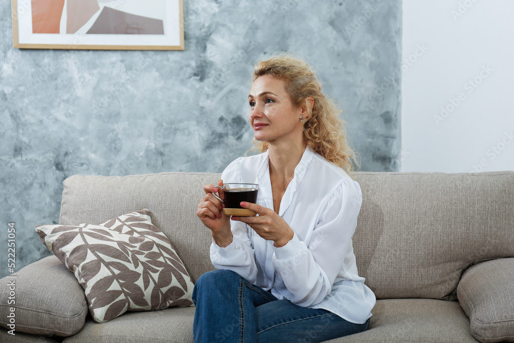 Portrait of adult beautiful woman wearing a white blouse sitting on the couch holding a big glass cup of black coffee. Copy space, background, close up