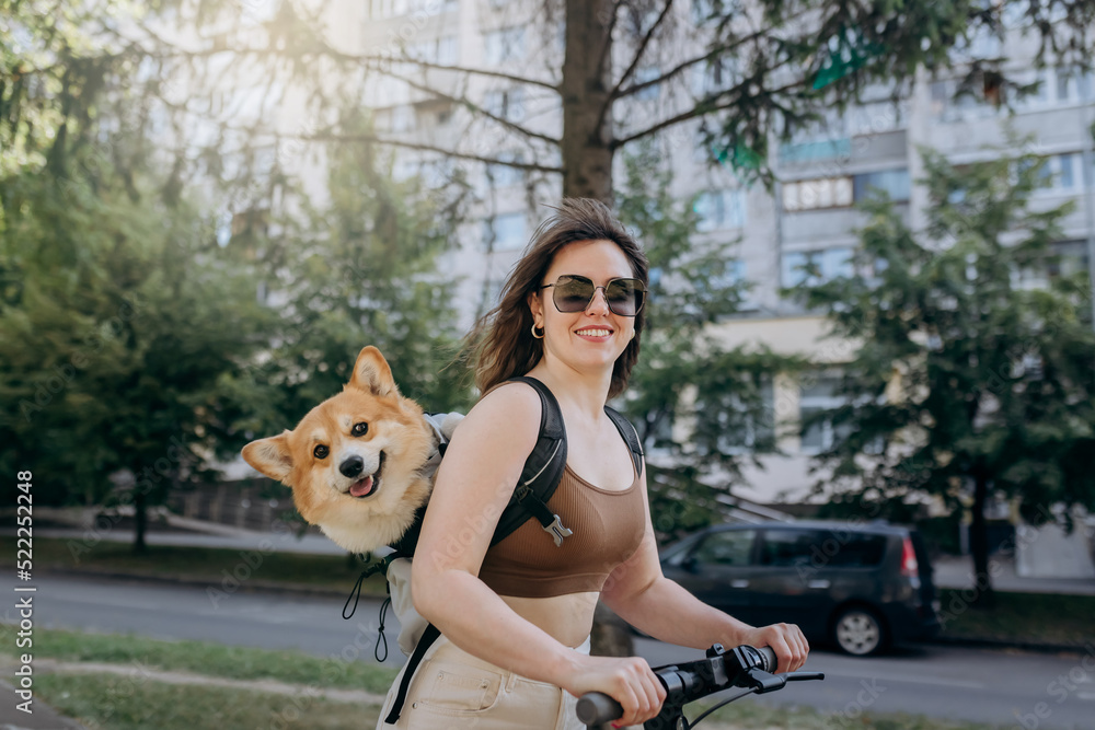 Happy smiling woman traveler is riding her electro scooter in city parkland with dog Welsh Corgi Pembroke in a special backpack Happy smiling woman traveler is riding her electro scooter in city parkland with dog Welsh Corgi Pembroke in a special backpack