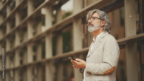 Profile mature businessman with beard in eyeglasses wearing gray jacket looking up at street signs and map trying to find her way using cellphone photo