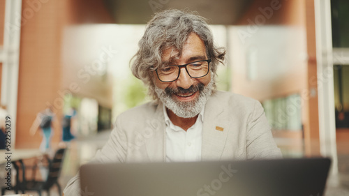 Mature businessman with beard in eyeglasses wearing gray jacket sits in cafe. Middle aged manager having video conference, video call on laptop pc computer sit at cafe outdoor