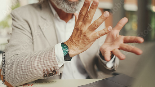 Close-up of the hands of mature businessman having video conference, video call on laptop pc computer sit at cafe outdoor. Middle aged manager massage hands, gesture