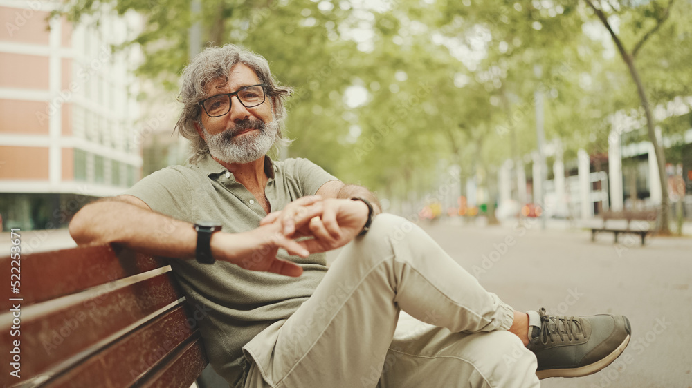 Obraz premium Thoughtful middle-aged man with gray hair and beard wearing casual clothes sits on bench. Mature gentleman in eyeglasses turns his head and looks into the camera