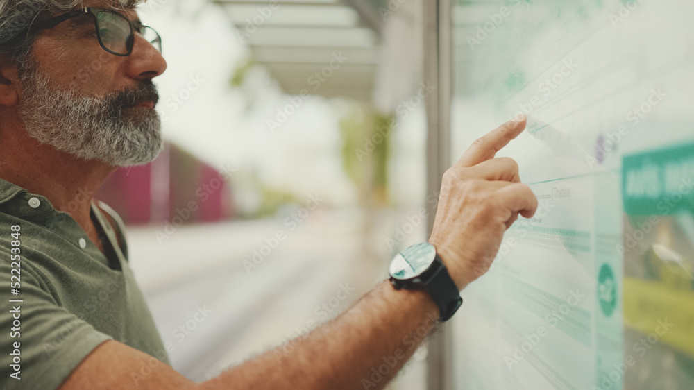 Clouse-up, profile of middle-aged man with gray hair and beard, runs his finger along the city transport schedule. Mature gentleman in eyeglasses is waiting for tram Clouse-up, profile of middle-aged man with gray hair and beard, runs his finger along the city transport schedule. Mature gentleman in eyeglasses is waiting for tram